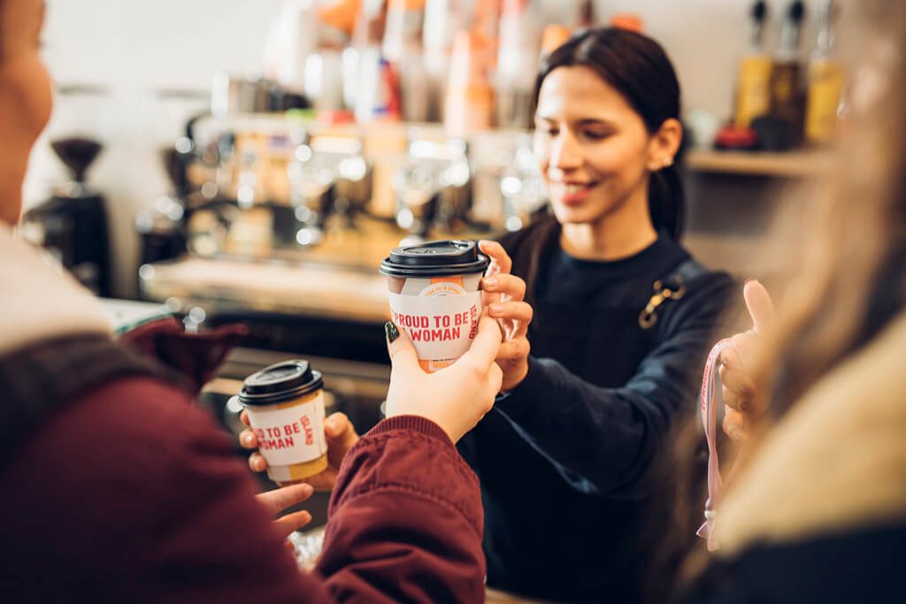 Coffee Island Baristi offering coffee to customers with International Women's Day sleeve