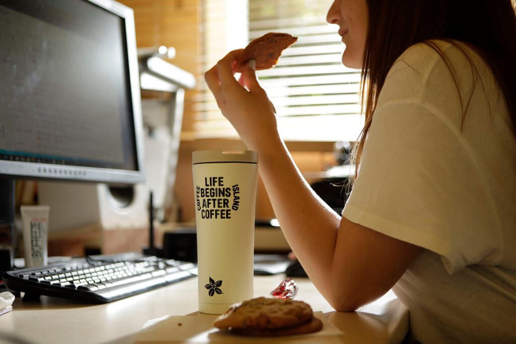 A WOMAN WORKING AND EATING COFFEE ISLAND'S COOKIES AND ENJOYING HER COFFEE IN A COFFEE ISLAND THERMOS.